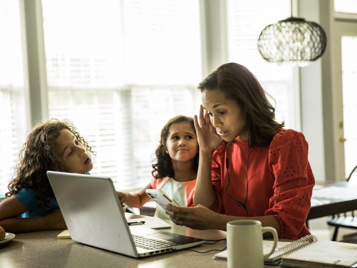 Mother looks stressed, sitting at a table with a computer and cell phone while working from home. Her two young children are talking to her and she sits with a hand on her head.