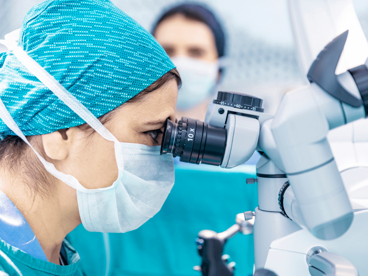 doctor wearing protective face mask and surgical cap, looking into the eye piece of a robotic surgery device.