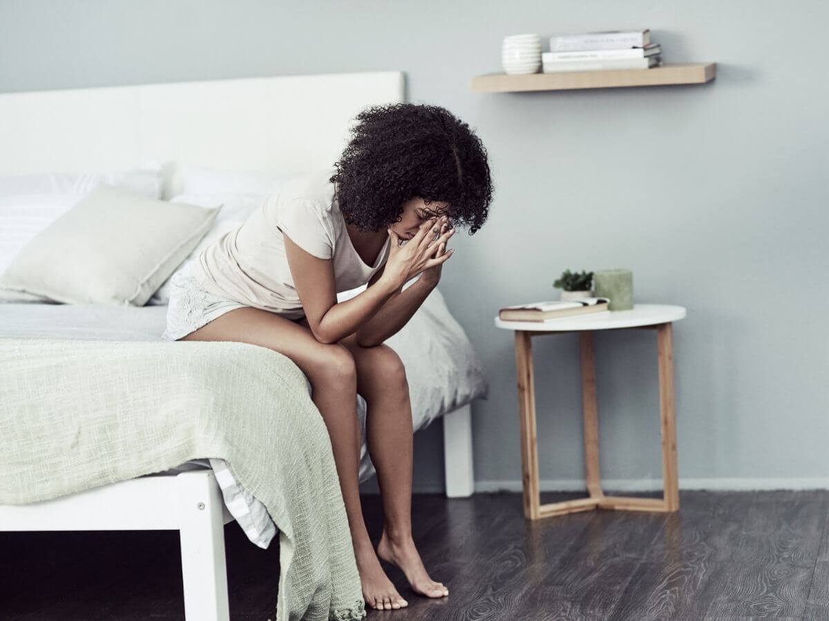 Woman sits on the edge of the bed with her head in her hands.