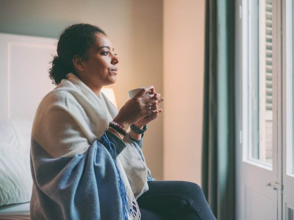 Woman sitting under a blanket holding a cup of coffee looking out the window