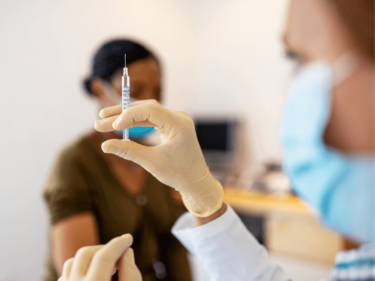 Nurse in foreground prepares a syringe, patient, out of focus in background waits to get vaccinated.