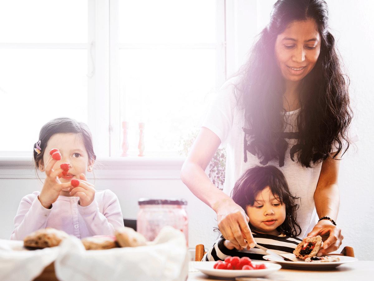 Mother and 2 young daughters eating breakfast, mother helping the youngest