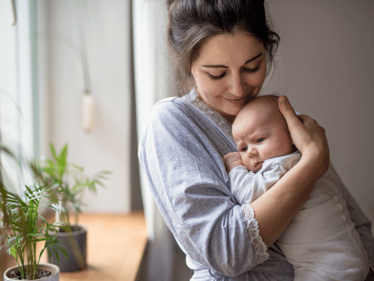 Mother stands near a window holding her newborn baby on her chest