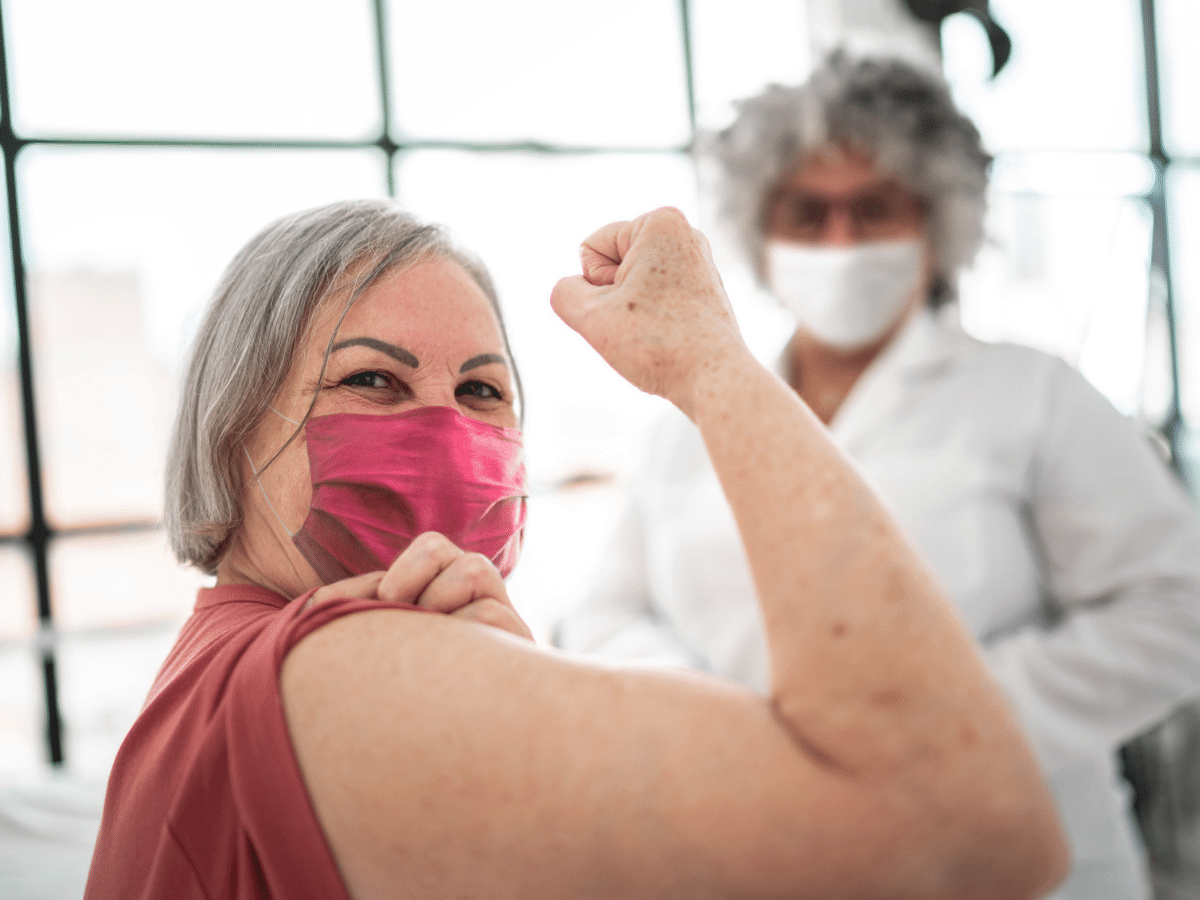 Older woman showing her arm at vaccine appointment with nurse visible in the background